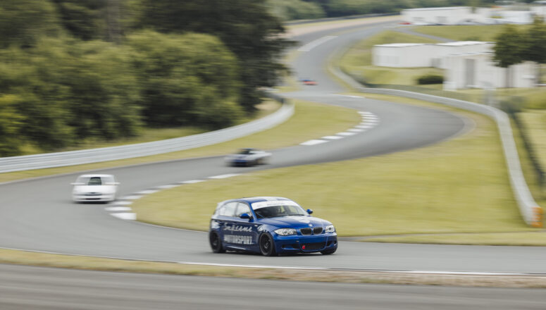 Blauwe BMW E87 130i rijdt op hoge snelheid op het circuit van Bilster Berg. Het circuit maakt een slalom bocht en is omringd door groen gras en bos.