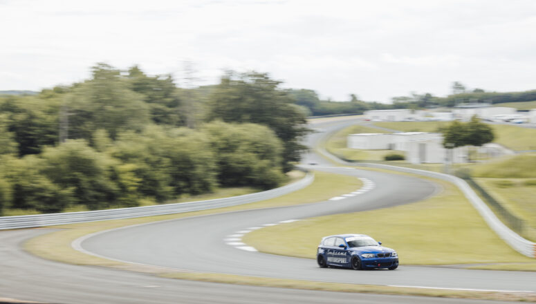 BMW 130i Le Mans Racer Blauwe BMW E87 130i rijdt op hoge snelheid op het circuit van Bilster Berg. Het circuit maakt een slalom bocht en is omringd door groen gras en bos.