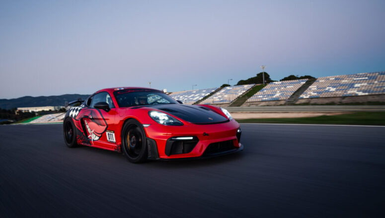 Een rode sportwagen met een zwarte motorkap en racingdecoratie rijdt met hoge snelheid over een racecircuit bij zonsondergang. Op de achtergrond zijn tribunes met blauwe en witte stoelen en lage heuvels te zien.