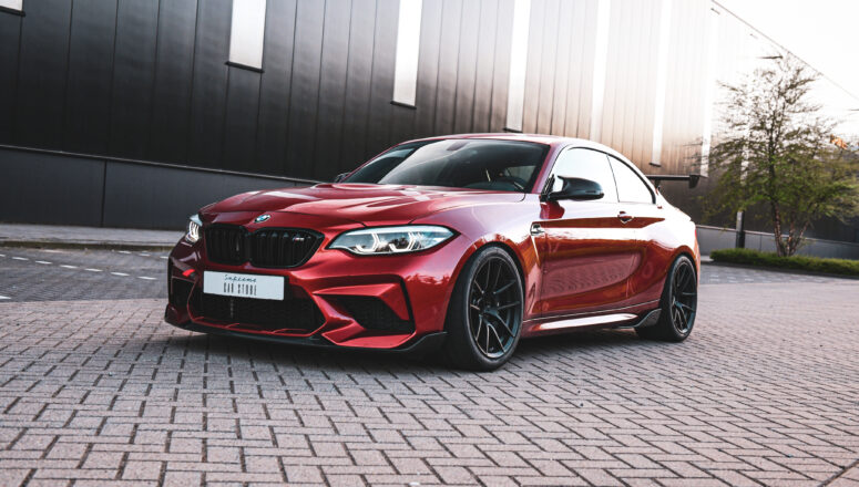 M2 Comp Oranje raceauto met zwarte wielen en spoiler. De auto is van het merk BMW en het model is een M2 Competition.