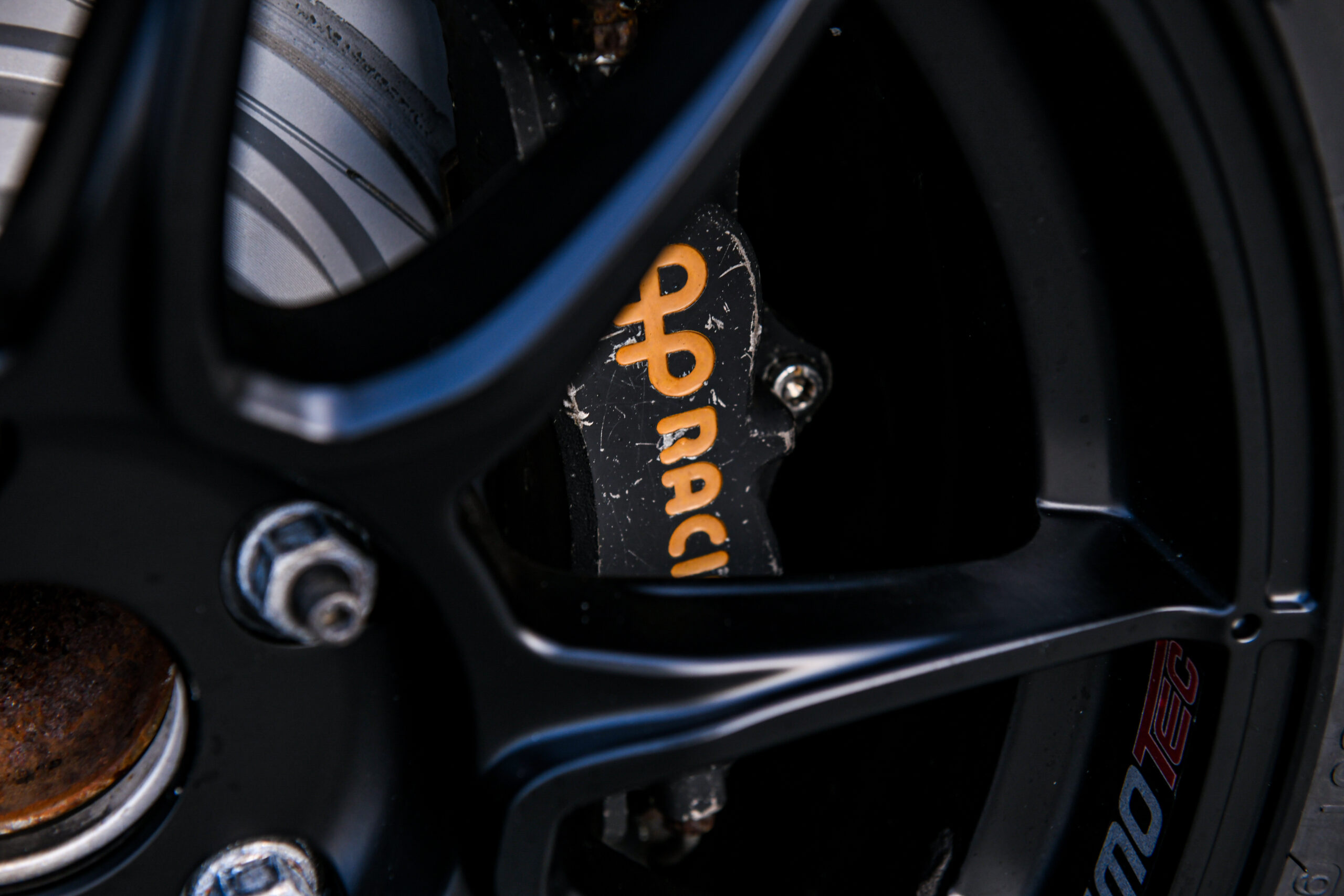 Ap Racing Brakes Zwarte remklauw met geel logo (Ap Racing) op de klauw. Deze is te zien langs de spaken van de zwarte wielen van de auto.